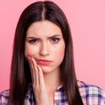 Brunette woman in a plaid shirt cringes and frowns because of her braces pain
