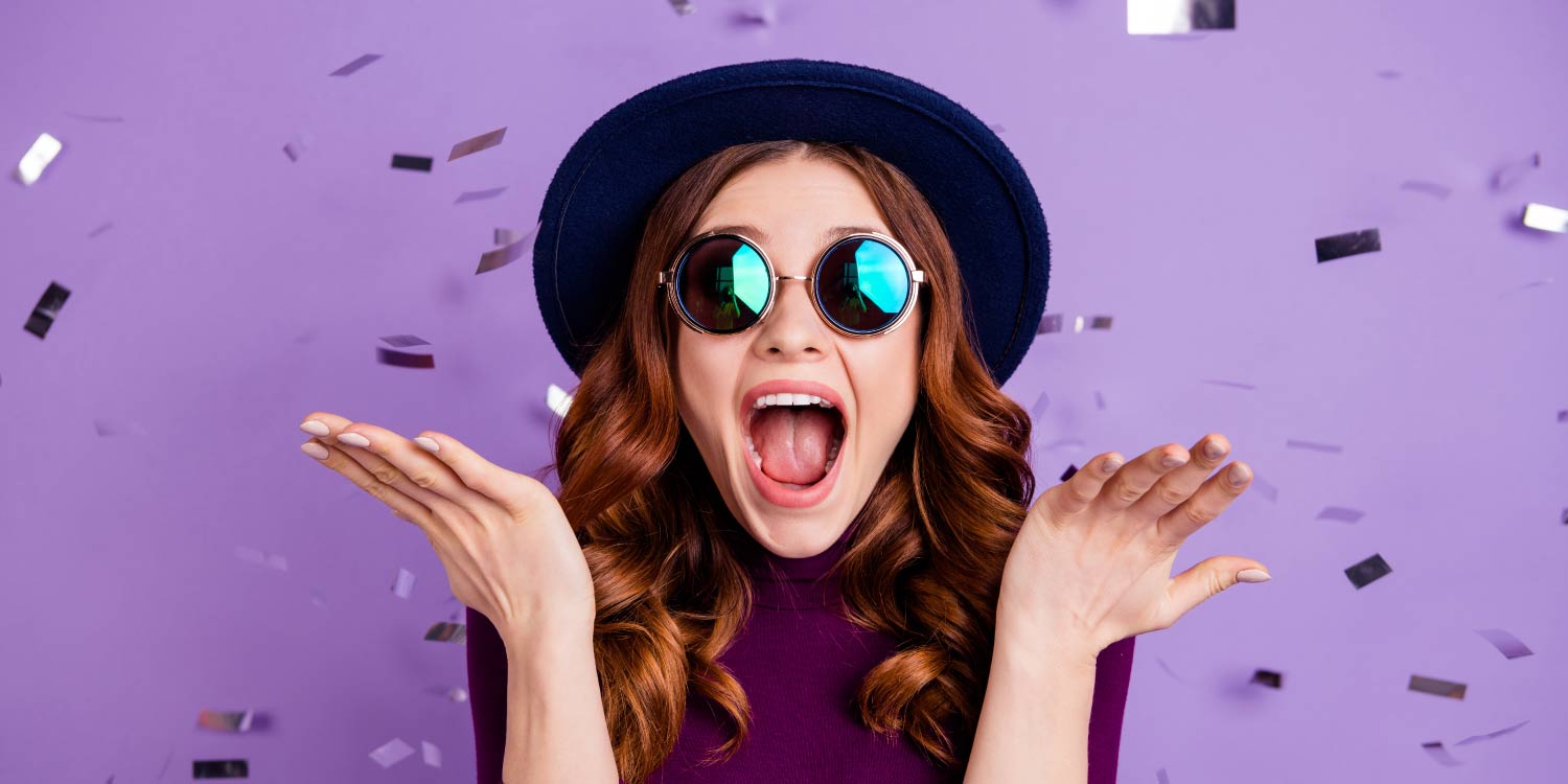 Brunette woman in a black hat and sunglasses celebrates with purple confetti getting her braces off