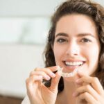brunette woman smiles as she puts in her Invisalign clear aligners to fix her crooked teeth