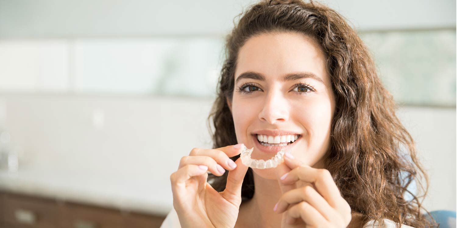 brunette woman smiles as she puts in her Invisalign clear aligners to fix her crooked teeth