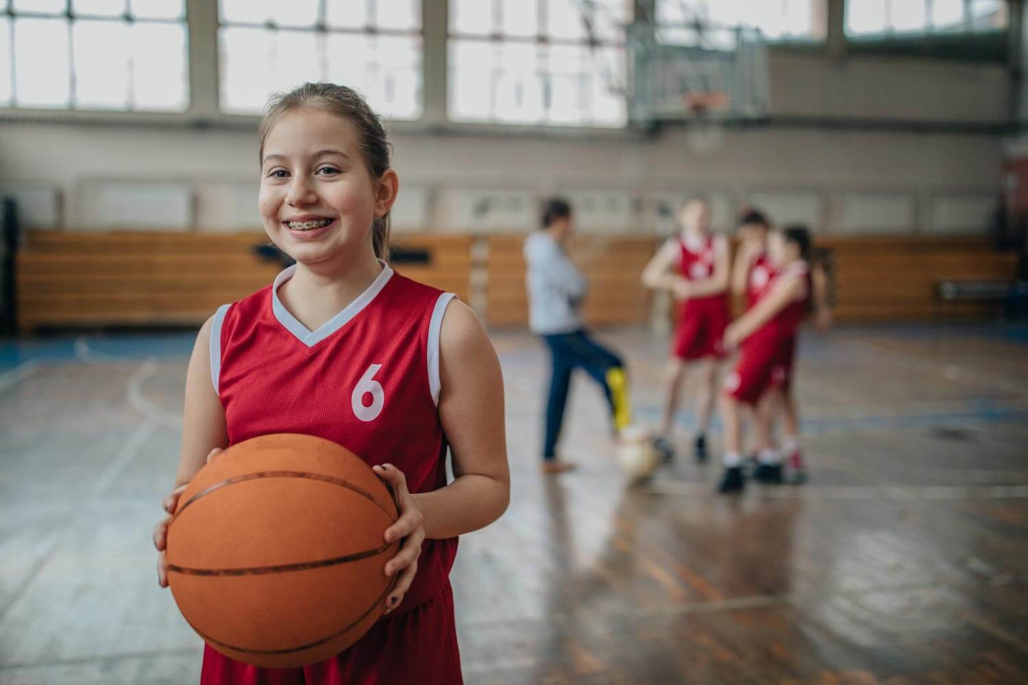 Young girl in a red jersey holds a basketball while wearing braces inside on a basketball court