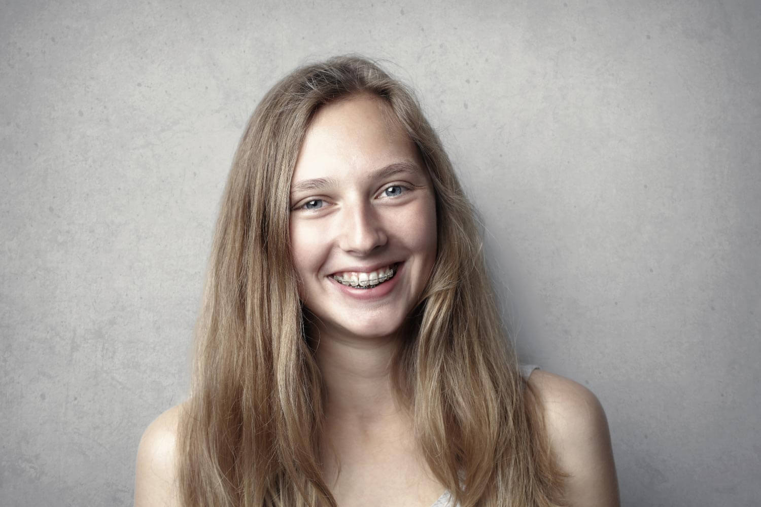 Brunette girl smiles with ceramic braces against a gray wall in Weatherford, TX
