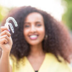 Blurred Black woman with big curly hair in a yellow blouse smiles while holding 2 Invisalign aligners outside on a walkway by some trees