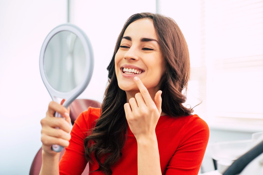 Brunette woman in a red blouse smiles at her straight smile in a handheld mirror