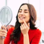 Brunette woman in a red blouse smiles at her straight smile in a handheld mirror
