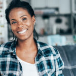 Black woman in a white shirt and a teal checkered flannel smiles with her braces while sitting on the couch