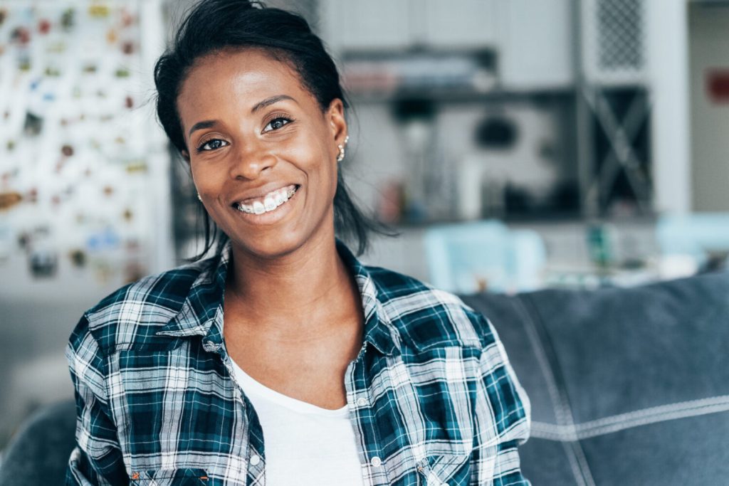 Black woman in a white shirt and a teal checkered flannel smiles with her braces while sitting on the couch
